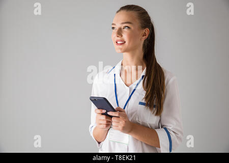 Foto von Eine schöne glückliche junge Frau cosmetologist Arzt über graue Wand Hintergrund holding Handy getrennt durch. Stockfoto