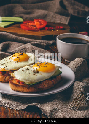 Italienische Toasts mit Gemüse und Spiegeleier auf weißen Teller und Tasse Kaffee über Grau grobes Tuch. Stockfoto