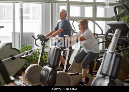 Ältere Menschen biken im Fitnessstudio. Ältere Paare tun, indoor Radfahren im Fitnesscenter. Fitnessstudio Training Konzept. Stockfoto