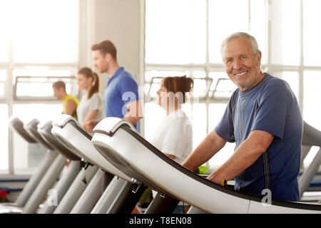 Lächelnd alter Mann trainieren im Fitnessstudio. Ältere Menschen in Sportkleidung auf Laufband und Kamera. Bleiben Sie aktiv durch ihre Jahre. Stockfoto