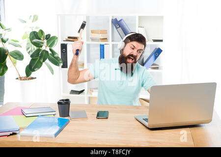 Hasse office Routine. Man bärtige Kerl Kopfhörer Büro schwingen Hammer auf dem Computer. Langsame Internetverbindung. Veraltete Software. Computer lag. Gründe für Computer zurückbleiben. Wie fix langsam hinterher. Stockfoto