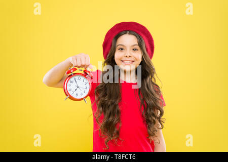 Shopping, Verkauf und Zeit Konzept. Pariser Kind auf Gelb. Kind mit Wecker. Zeitlose Mode. glückliches Mädchen mit langen Locken in Baskenmütze. Schönheit Friseur. Kleines Mädchen im französischen Stil hat. Stockfoto