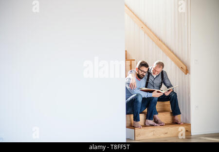 Ein erwachsener Sohn und Vater auf Treppen im Innenbereich, zu Hause zu sitzen, auf der Suche nach Fotos. Stockfoto
