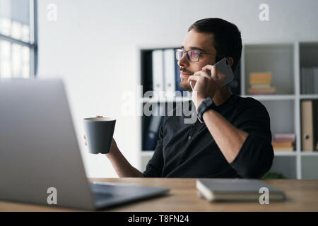 Junger Mann mit Handy und Computer, Empfangen von Anrufen, im Gespräch mit Partnern Stockfoto