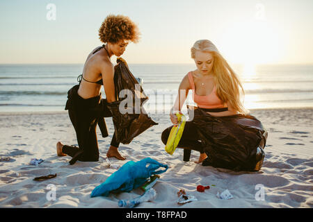 Zwei junge Frauen, die Kommissionierung, Abfall vom Strand. Weibliche surfer Reinigung der Strand, der Erfassung der Wurf in den Müllsack. Stockfoto