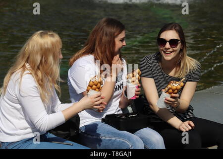 Moskau, Russland - Mai 2019: Drei glückliche Mädchen in der Nähe von einem Brunnen sitzen und Waffeln mit Eis im sonnigen Tag essen. Konzept der kalorienahrung Stockfoto