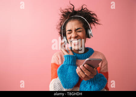 Bild der emotionalen Schöne junge afrikanische Frau posiert über rosa Wand Hintergrund Musik hören mit Kopfhörern über Mobiltelefon isoliert. Stockfoto