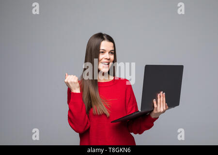 Portrait von überglücklich Mädchen im roten Pullover ballte die Faust im Sieger Geste halten Laptop, auf grauem Hintergrund Stockfoto