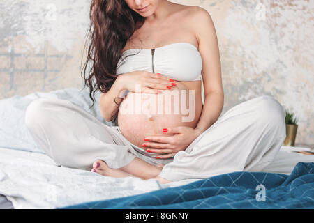 Foto zuschneiden der schönen schwangeren Mädchen Junge Frau mit langen, lockigen Haar sitzt im Lotussitz auf dem Bett. Schwangerschaft Yoga und Natur ethnischen Schönheit Konzept Stockfoto