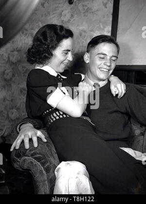 1950, historische, ein glückliches junges Paar in Liebe ..... ein junger Mann sitzt drinnen auf einem Sessel mit seiner Freundin auf seinem Schoß und mit ihre Arme um ihn, Manchester, England, UK. Stockfoto