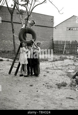 1970 s, historische, zum Aufhängen von einem reifen Schwingen.... vier junge Kinder zusammen draußen stehen in einem sandigen Industriegebiet von Land oder Hof, das Halten eines Gummi Reifen, während ein junges Mädchen in einer Leiter kletterte, um einen kleinen Baum, versucht sie es mit String zu befestigen. Trye Schaukeln sind ein großer Spaß für Kinder die spielen beim Aufwachsen und es ist auch das Recycling. Stockfoto