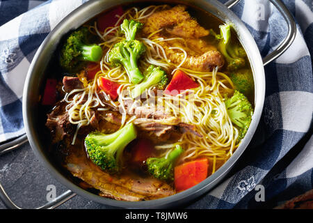Huhn Nudelsuppe mit Brokkoli, Karotten und Gewürze in einem Metall Kochtopf mit Löffel, Küche Handtuch und Baguette in Scheiben geschnitten auf einer konkreten Tabelle, Schließen - Stockfoto