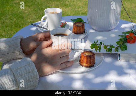 Erholsame Zeit an einem sonnigen Tag im Garten. Genießen Sie Kaffee mit caneles. Anordnung mit Händen und einer Frau eine weiße serviert. Stockfoto