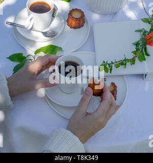 Genießen Kaffee Zeit im Garten. Serviert mit Kaffee, canele, Blumen und Buch im Garten. Anordnung mit den Händen einer Frau. Stockfoto