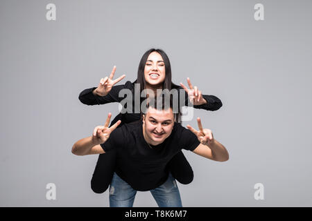 Junge lustig Paar, Freund piggy backing Freundin, Mädchen, Frieden Zeichen mit den Händen über grauer Hintergrund Stockfoto