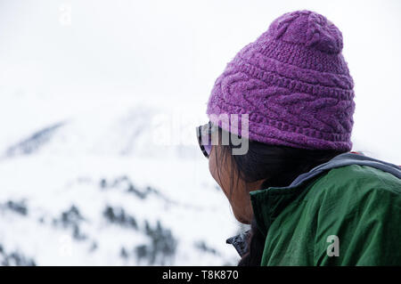 Asiatische Frau in lila Hut sieht nach oben in Richtung des verschneiten Berg bei bedecktem Himmel Winter Tag Stockfoto