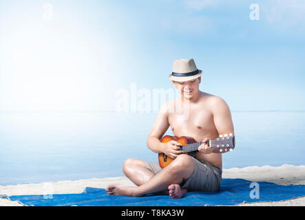 Asiatischer Mann in Hut sitzen und Gitarre Spielen auf dem Teppich in Strand. Sommer Urlaub Stockfoto