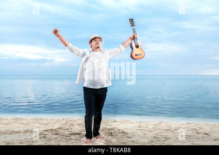 Asiatische Geschäftsmann in Hut und weiße Kleidung hob beide Hände halten Gitarre am Strand. Sommer Urlaub Stockfoto