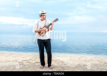 Asiatische Geschäftsmann in Hut und weiße Kleidung Gitarre spielen am Strand. Sommer Urlaub Stockfoto