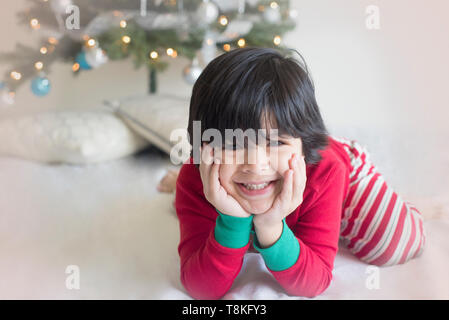 Hübsche Junge sitzt mit einem Weihnachtsbaum im Schlafanzug Stockfoto