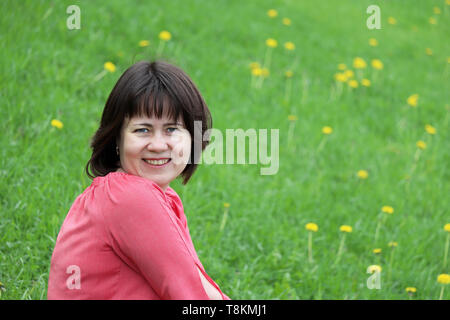 Gerne Frau mittleren Alters in rosa Kleid sitzt auf einer grünen Wiese mit Löwenzahn Blumen. Konzept von Glück, positive Emotionen, Freude, Ökologie, Freizeit Stockfoto