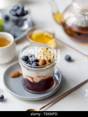 Frische, gesunde Frühstück mit Müsli mit Marmelade und Joghurt im Glas mit Blaubeeren, mit Tee und Honig in der Nähe von auf weißem Hintergrund Stockfoto