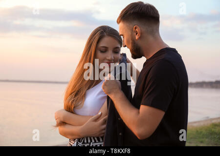 Junger Mann das Hemd auf seiner Freundin in der Nähe von Fluss bei Sonnenuntergang Stockfoto