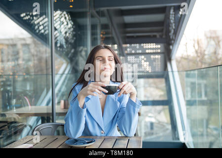 Die Liebe zum Kaffee. Portrait von süße Mädchen trinken Ihren Kaffee auf dem Balkon und über Terrasse mit Green Bush Hintergrund, trägt blaue Anzug, wh Stockfoto