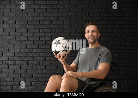 Junger Mann mit Kugel Sitzen im Rollstuhl gegen dunkle Mauer Stockfoto