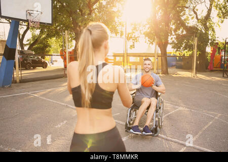 Junger Mann im Rollstuhl und sportliche Frau Training mit Ball im Freien Stockfoto