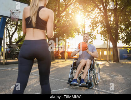 Junger Mann im Rollstuhl und sportliche Frau Training mit Ball im Freien Stockfoto