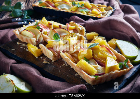 In der Nähe der frisch gebackene Ananas und Apfel süße Torte auf einem Schneidebrett in Scheiben geschnitten auf einem alten Holztisch mit braunem Stoff, horizontale Ansicht von abov Stockfoto