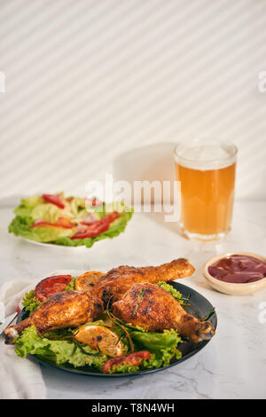 Gegrillte Hähnchenschenkel gebraten auf den Grill auf dunklen Platte mit Tomatensauce in eine Schüssel und Salatblätter, Glas Becher Bier Stockfoto