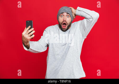 Bild der kaukasischen Mann 30 s Kopf greifen und halten Smartphone beim Stehen über den roten Hintergrund isoliert Stockfoto