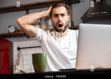 Junger Mann mit Laptop Computer, während am Küchentisch sitzen Schockiert Stockfoto