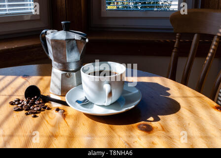 Aluminium mocha Coffee Pot, weiße Tasse mit schwarzem Kaffee, Untertasse und silbernen Löffel und Kaffeebohnen auf einem Sonnenverwöhnten Küche Tabelle Stockfoto