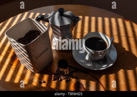 Weiß Kaffee Dose mit Kaffeebohnen, Aluminium mocha Coffee Pot, weiße Tasse mit schwarzem Kaffee, Untertasse und silbernen Löffel und Kaffeebohnen am Küchentisch Stockfoto