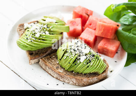 Scheiben Avocado mit weichen, weißen Käse auf Toast mit Wassermelone Eiswürfel serviert. Gesund Sommer Frühstück oder Mittagessen Stockfoto