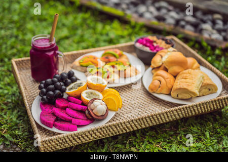 Frühstück auf einem Tablett mit Obst, Brötchen, Avocado Sandwiches, smoothie Schüssel stehen auf dem Gras Stockfoto