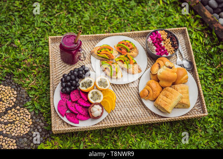 Frühstück auf einem Tablett mit Obst, Brötchen, Avocado Sandwiches, smoothie Schüssel stehen auf dem Gras Stockfoto