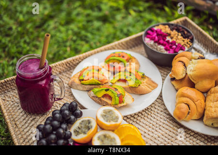 Frühstück auf einem Tablett mit Obst, Brötchen, Avocado Sandwiches, smoothie Schüssel stehen auf dem Gras Stockfoto