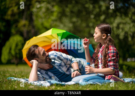 Vater und Tochter auf der blauen Decke mit Seifenblasen im Park. Bunten Regenschirm auf dem Hintergrund. Stockfoto