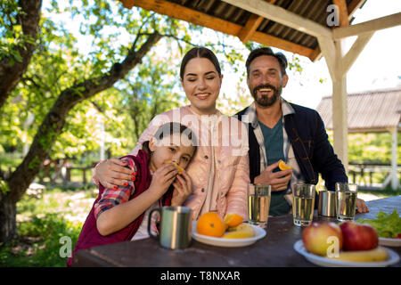Glückliche Familie Sitzen durch die Tabelle in den Park. Platten mit verschiedenen Früchten und Gläser mit Wasser sind auf dem Tisch. Stockfoto