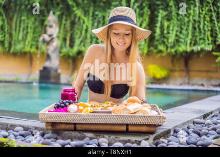 Junge Frau beim Frühstück auf einem Tablett mit Obst, Brötchen, Avocado Sandwiches, smoothie Schüssel am Pool. Sommer gesunde Ernährung, Veganes Frühstück. Lecker Stockfoto