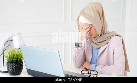Müde muslimischen Frau im hijab und traditionellen Verschleiß funktioniert und Typen auf Laptop, setzt sie ihre Brille aus und reibt ihre Augen in ihren Arbeitsplatz im Büro zu sitzen. Stockfoto