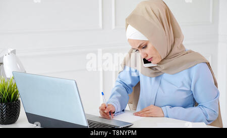 Junge muslimische Frau in Hijab spricht mit Client auf dem Handy erklären, Situation, Papier und Bildschirm auf Ihrem Arbeitsplatz im Büro. Stockfoto