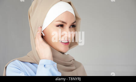 Portrait von muslimischen jungen Frau hijab tragen, beige und blau Kleid ist das drahtlose Headset im Ohr und sprechen das Smartphone mit Headset. Schnurlose Technologie. Stockfoto