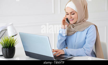 Porträt der jungen schönen muslimische Frau in beige Hijab und blauen Kleid arbeitet im Büro, Mobiltelefon und tippen auf Laptop an Ihrem Arbeitsplatz. Stockfoto