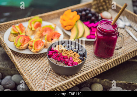 Frühstück auf einem Tablett mit Obst, Brötchen, Avocado Sandwiches, smoothie Schüssel am Pool. Sommer gesunde Ernährung, Veganes Frühstück. Lecker Ferienhäuser Konzept Stockfoto