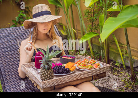 Junge Frau essen Frühstück in einem Sessel auf einem Tablett mit Obst, Brötchen, Avocado Sandwiches, smoothie Schüssel am Pool. Sommer gesunde Ernährung, vegan Stockfoto